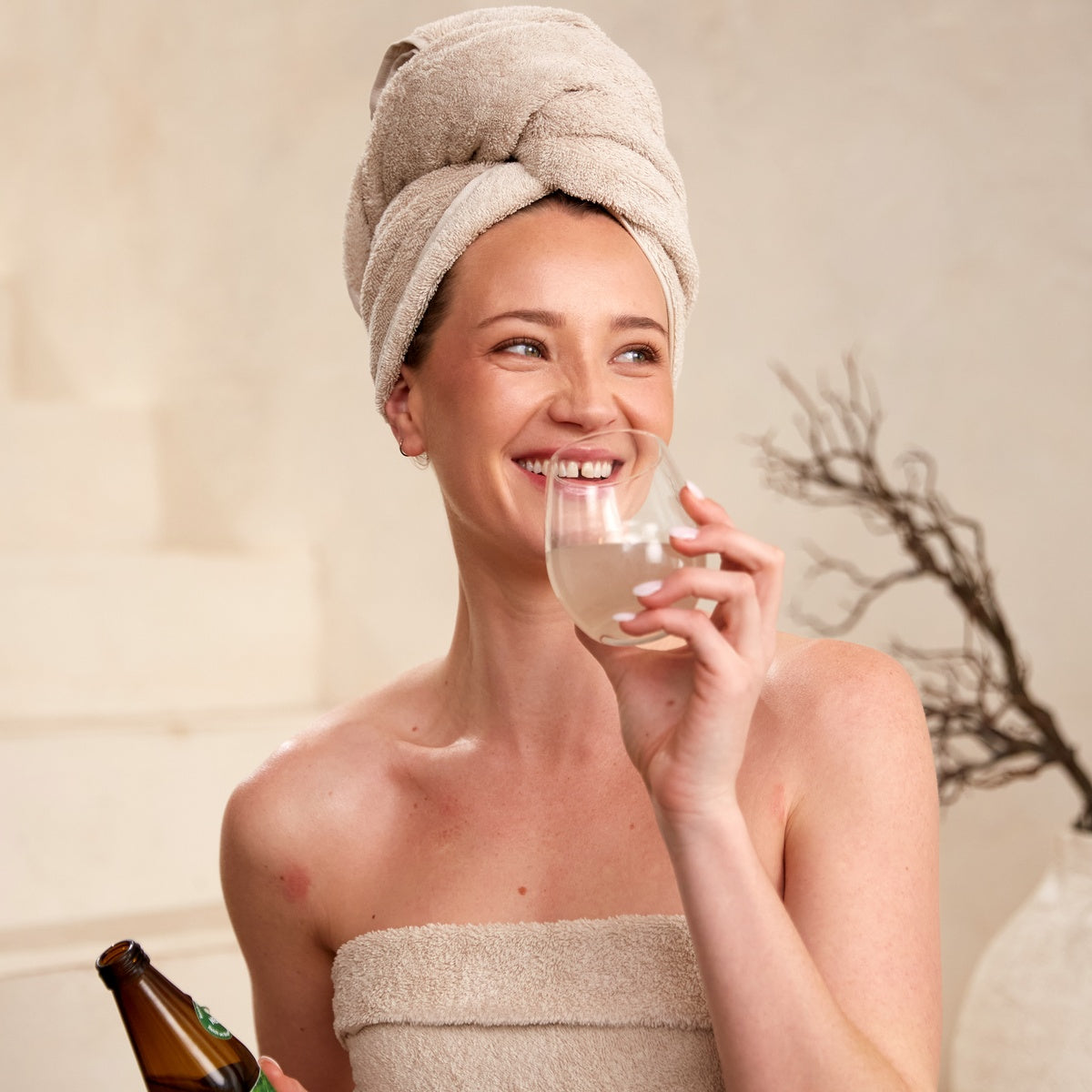 Lächelnde Frau mit Handtuch-Turban trinkt Aloe Vera BioUrsaft und hält Pharmos Natur Aloe Vera BioUrsaft Flasche